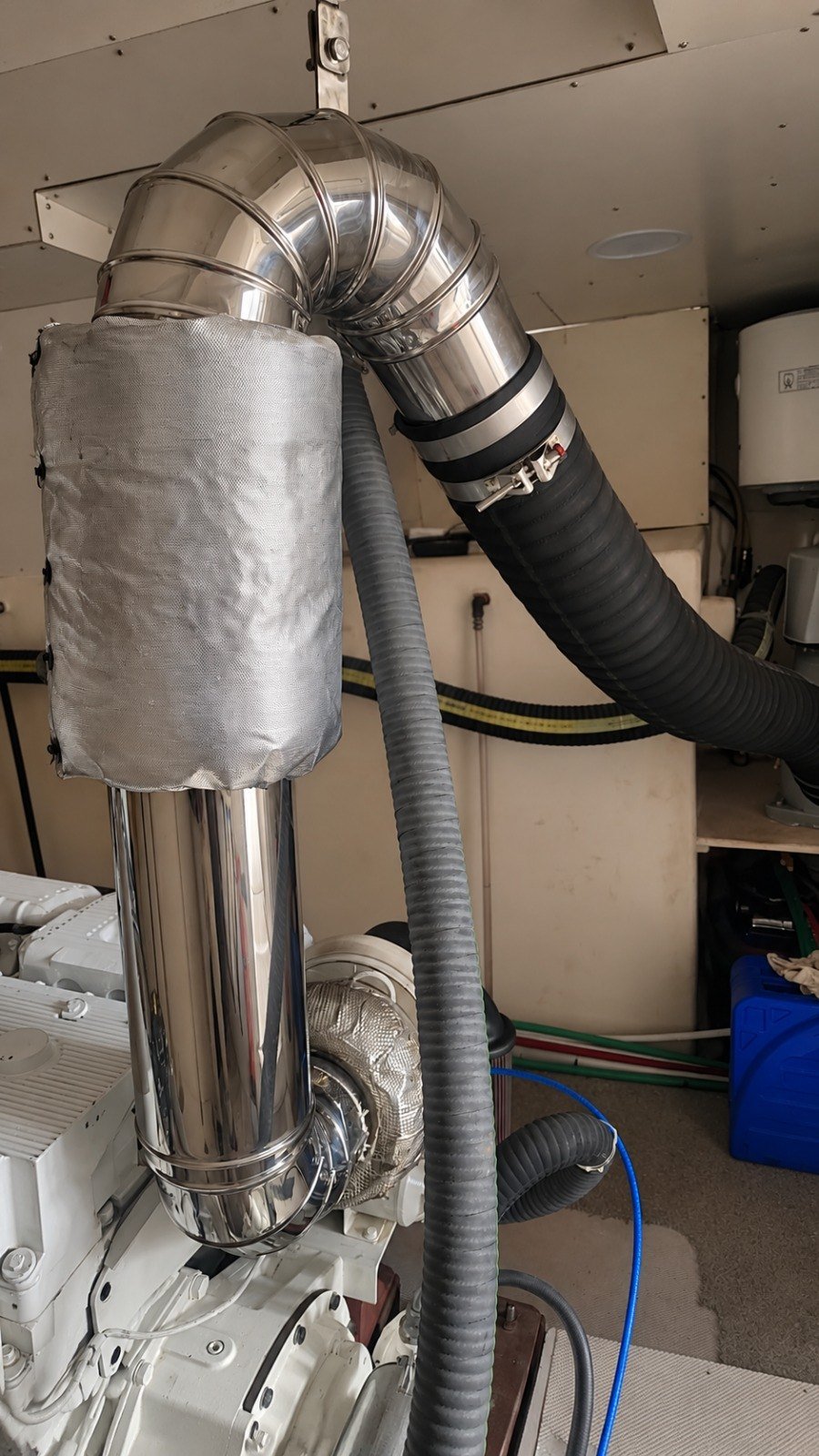 Polished stainless steel exhaust pipework with an insulated section and exhaust hose, rising from the engine in a vessel's engine room